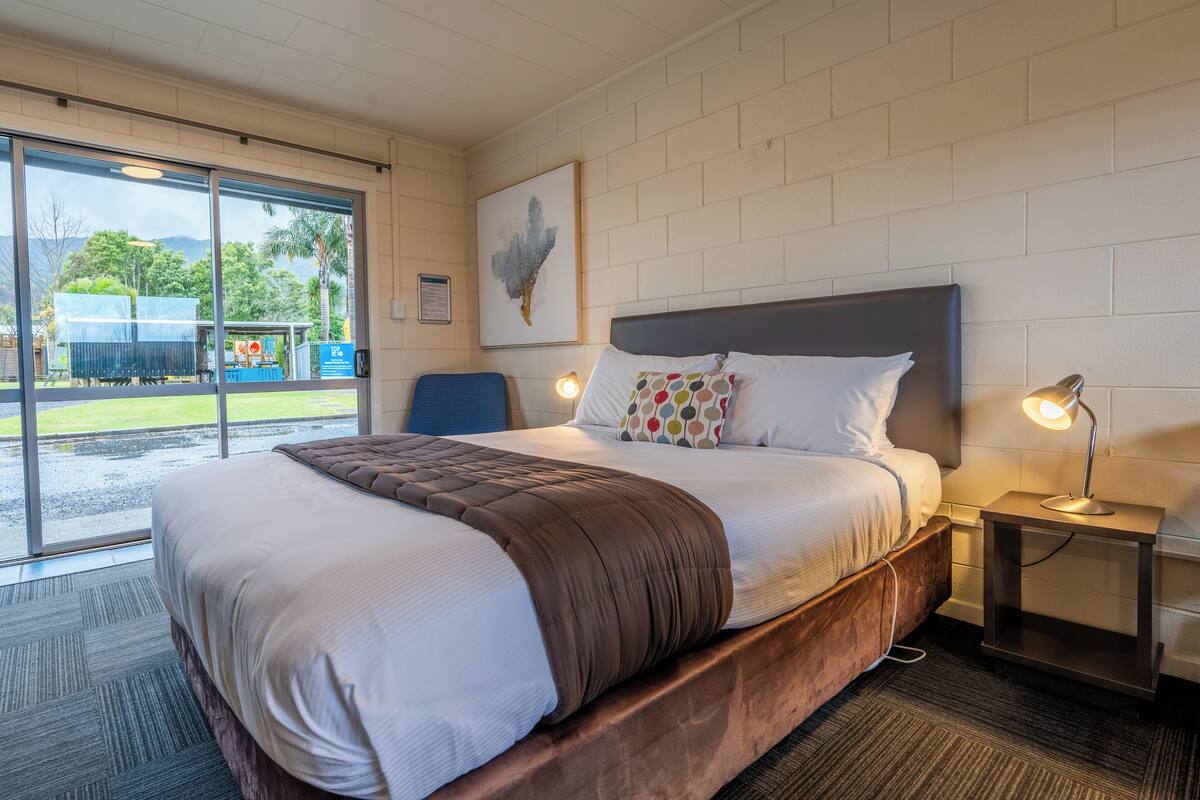 A hotel room with a large bed, a desk, and a chair. There is a window with a view of the outdoors.