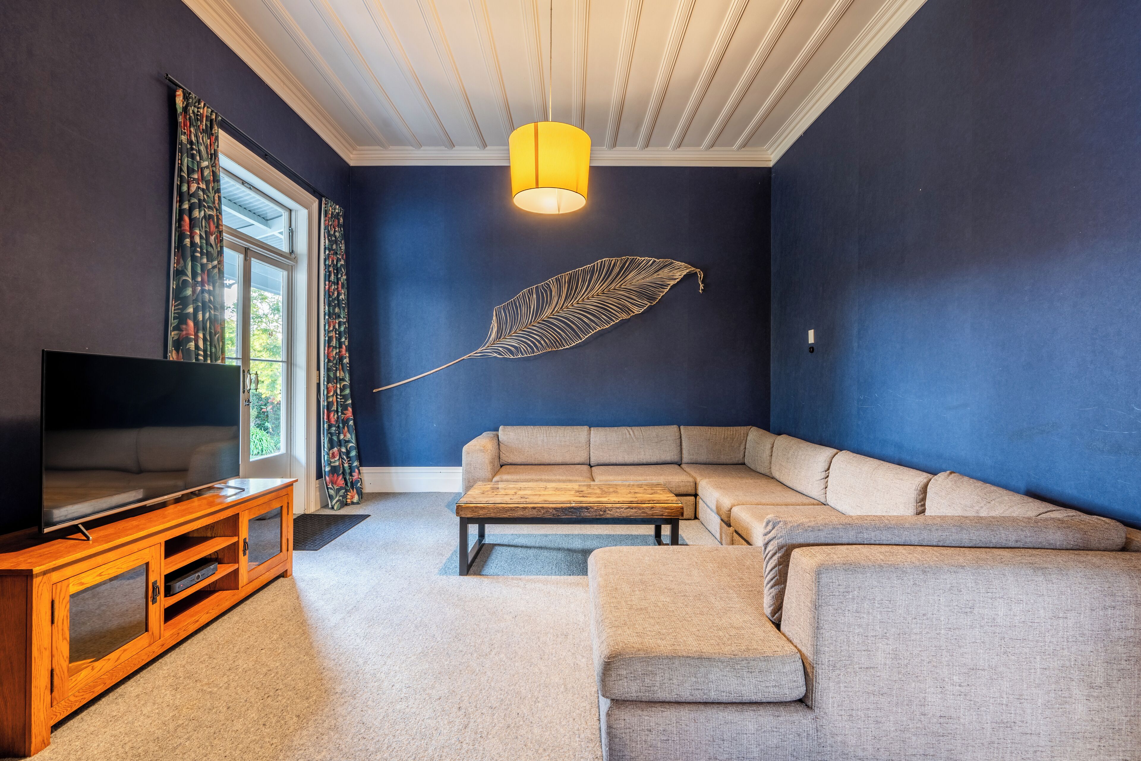 Ein Wohnzimmer mit einem großen Flachbildfernseher, einer Holz-Unterhaltungsecke, einem beigefarbenen Ecksofa und einem dekorativen Federwandbehang.
