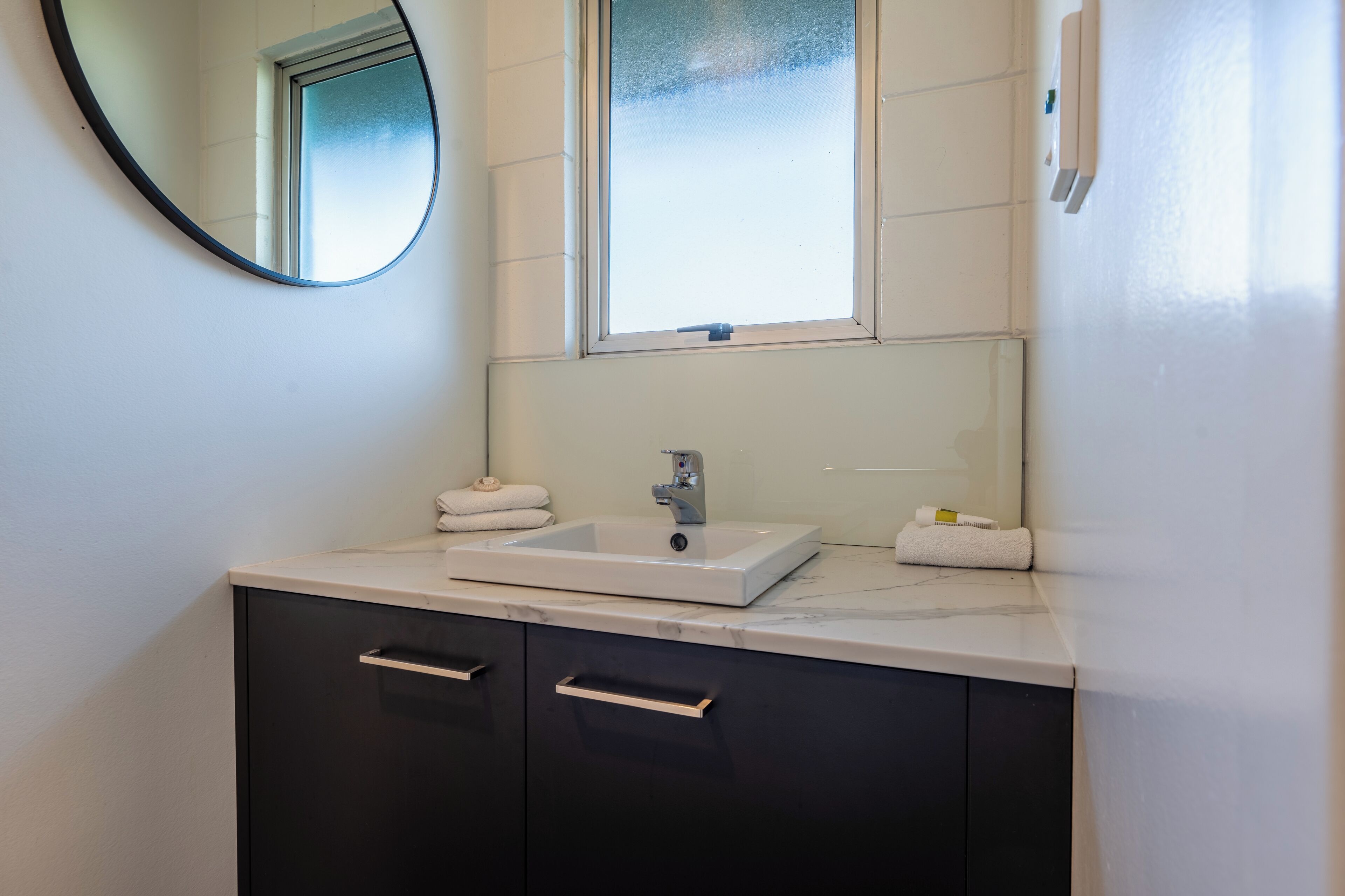 A bathroom with a white sink, dark cabinets, a mirror, and a window.