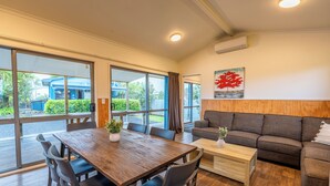 A modern living room with a wooden dining table, dark sofas, and large windows offering a view of the outdoors. - Tasman Holiday Parks – Coromandel (Coromandel)