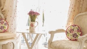 A room with a white chair, a small table with a vase of flowers, and a window with floral curtains. - Chigdem Hotel - Special Class (Cesme)