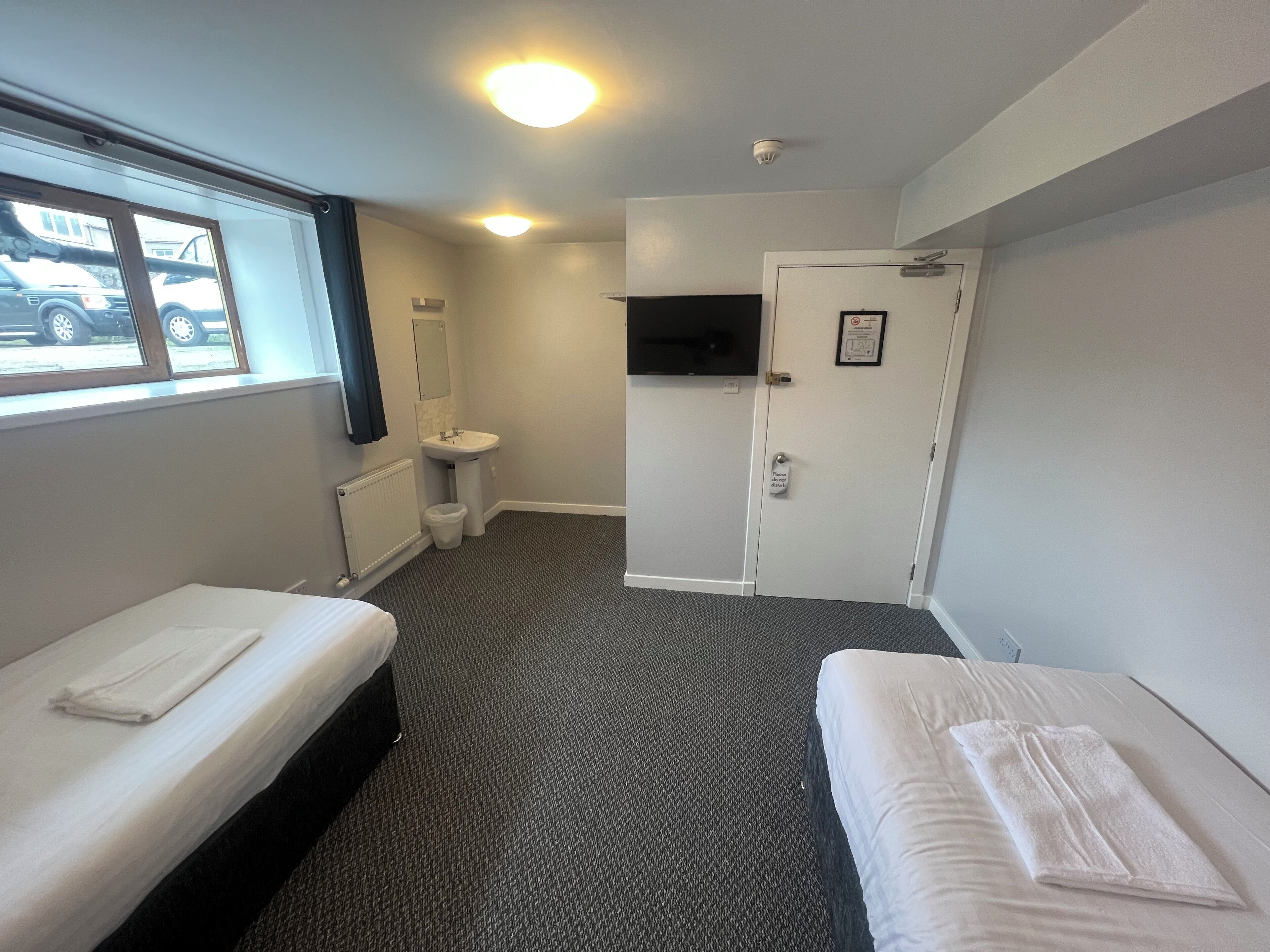 A hotel room with two single beds, a window with a view of parked cars, a sink, a toilet, and a door with a do-not-disturb sign.