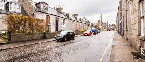 Une rue étroite bordée d’immeubles en pierre, de voitures garées, et où l’on aperçoit au loin le clocher d’une église.