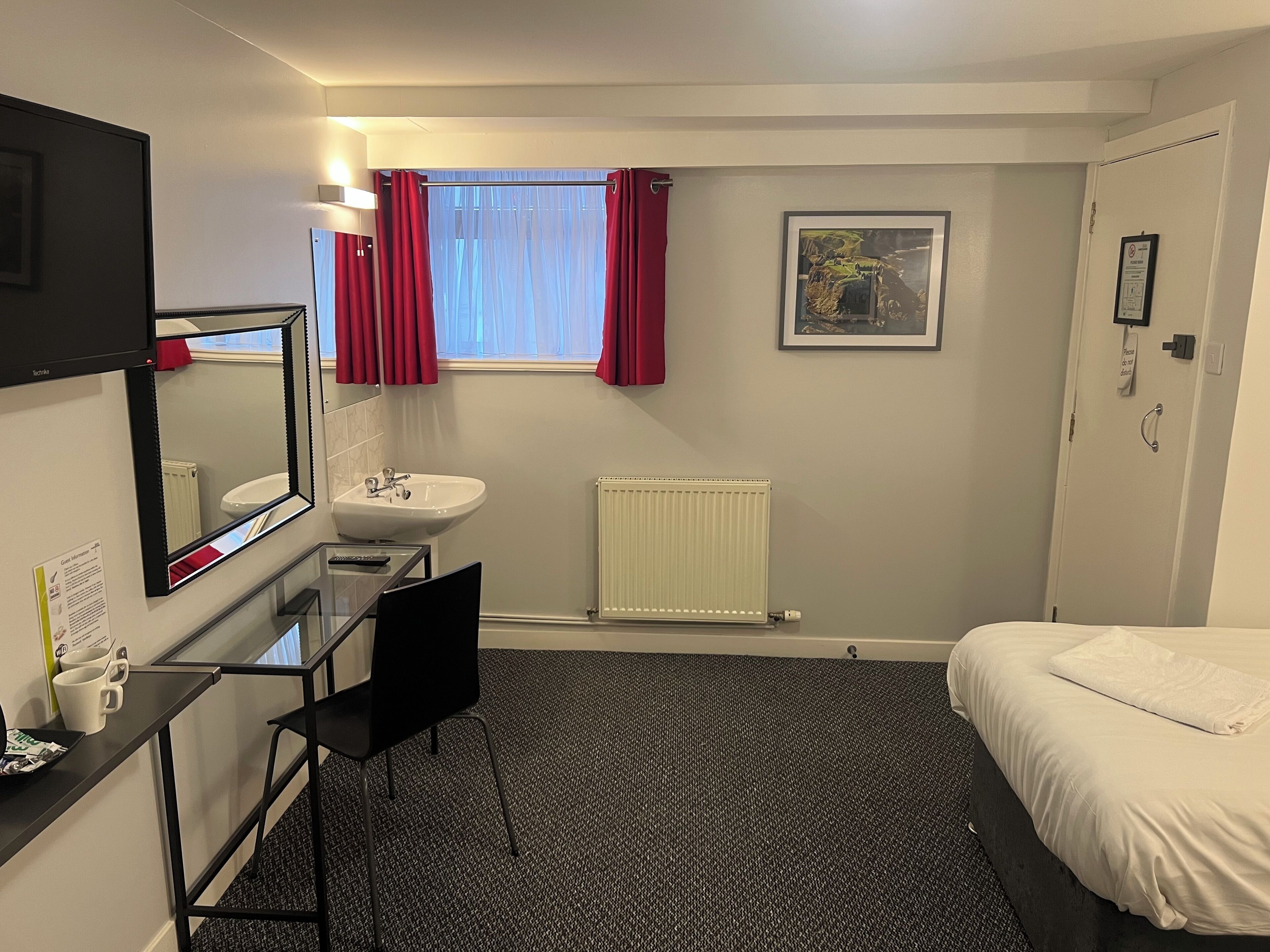 Une chambre d’hôtel comprenant un lit, un bureau avec une chaise, un lavabo, un miroir, une fenêtre avec des rideaux, un radiateur et un tableau encadré au mur.