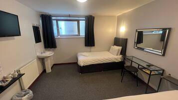 Habitación de hotel con cama, mesita de noche, espejo, ventana con cortinas, lavamanos y televisión en la pared.