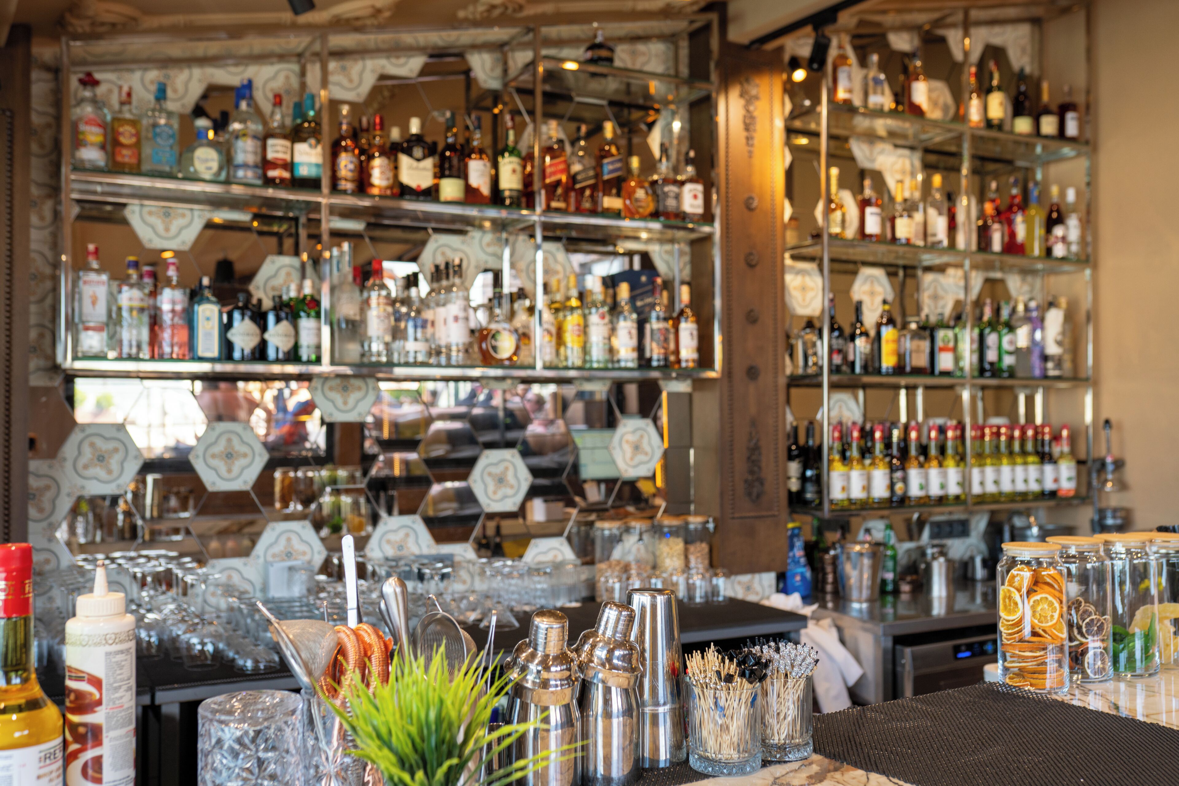 Un bar con estantes llenos de botellas de licor, una barra con vasos y botellas, y una planta decorativa.