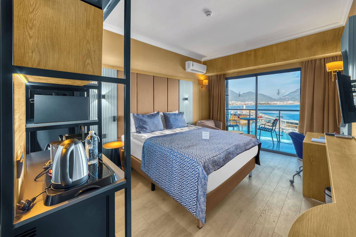 A hotel room with a bed, a desk with a kettle, a chair, and a view of the sea.