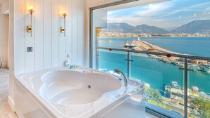 A modern bathroom with a large bathtub, a glass-enclosed balcony with a view of a coastal city and mountains. - Numa Port Hotel (Alanya)
