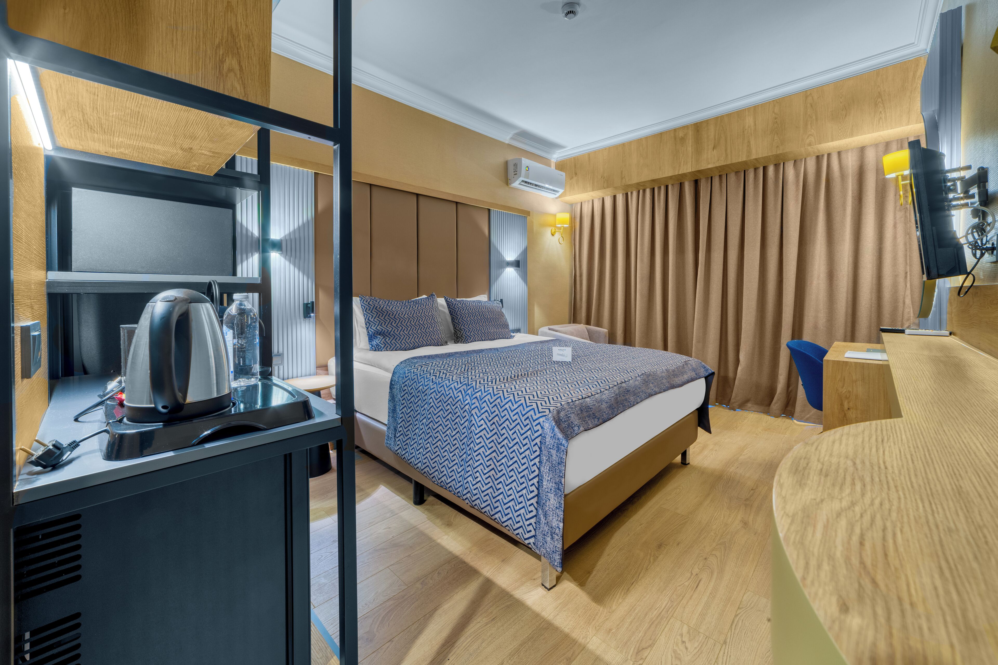 Moderni hotellihuone, jossa on sänky, yöpöytä ja työpöytä, sekä vedenkeitin ja vesipullo.