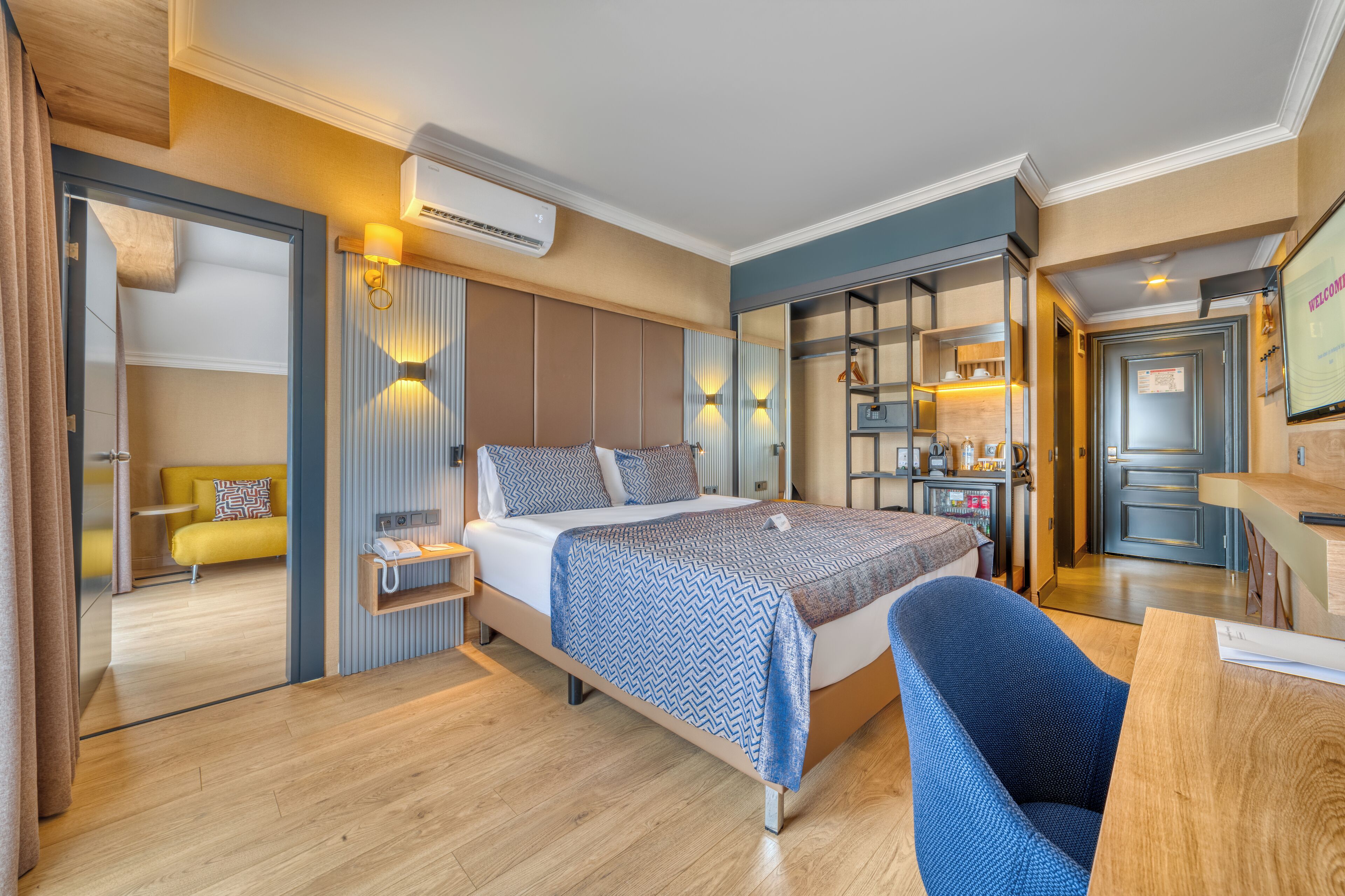 A modern hotel room with a bed, a desk, a chair, a small kitchenette, and a bathroom visible through an open door.