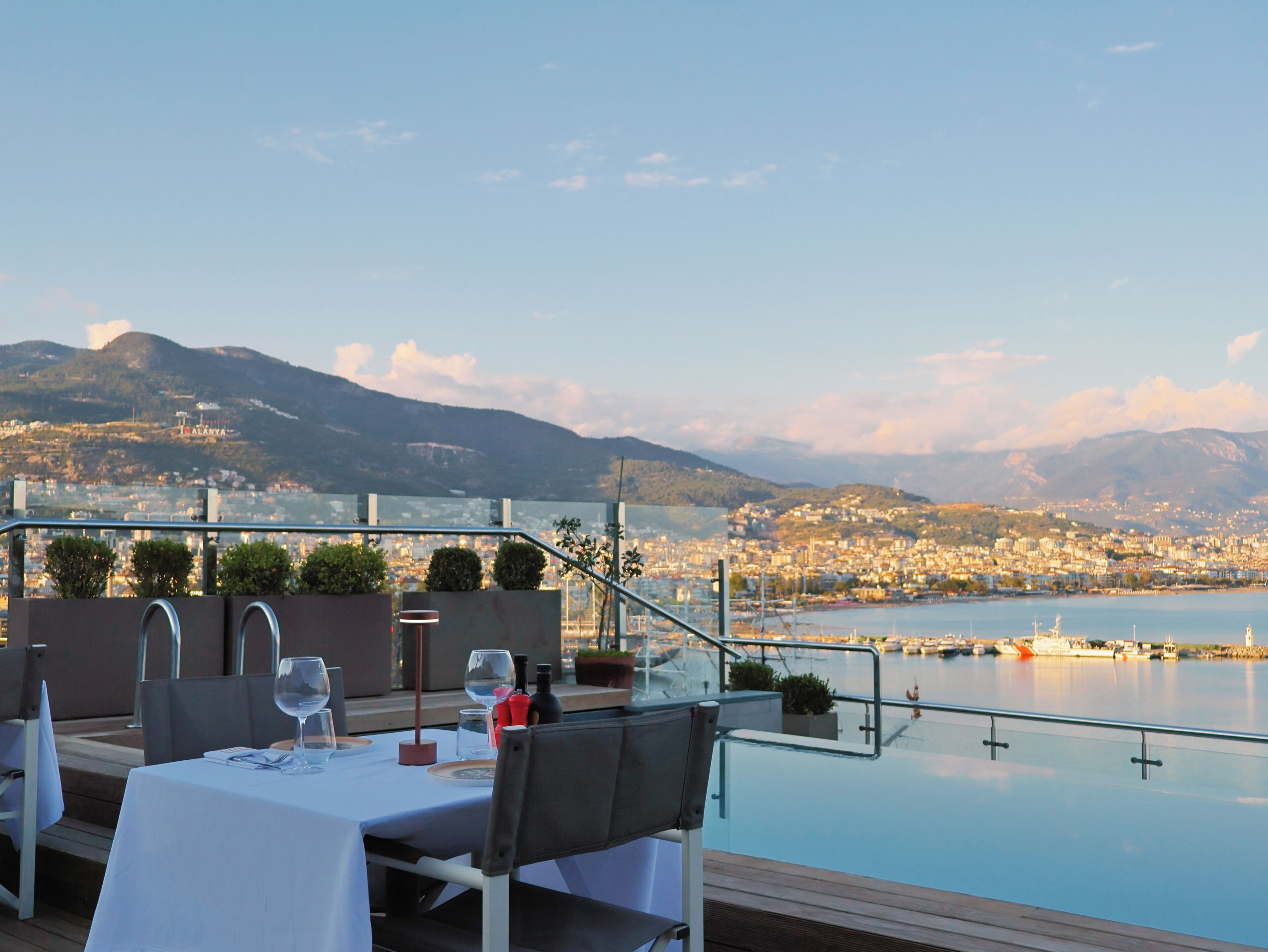 Une table à manger dressée avec une nappe blanche, des chaises, des verres à vin et une bouteille, donnant sur un paysage urbain au bord de l’eau, avec des montagnes en arrière-plan.