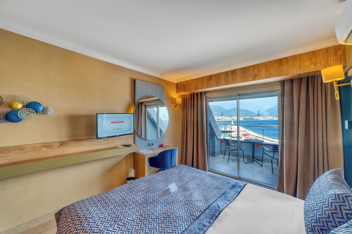 A hotel room with a bed, a desk, a chair, a TV, and a balcony with a view of the sea.