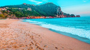 A beach with footprints leading to the water, a rocky cliff with buildings, and a clear sky. - Numa Port Hotel (Alanya)