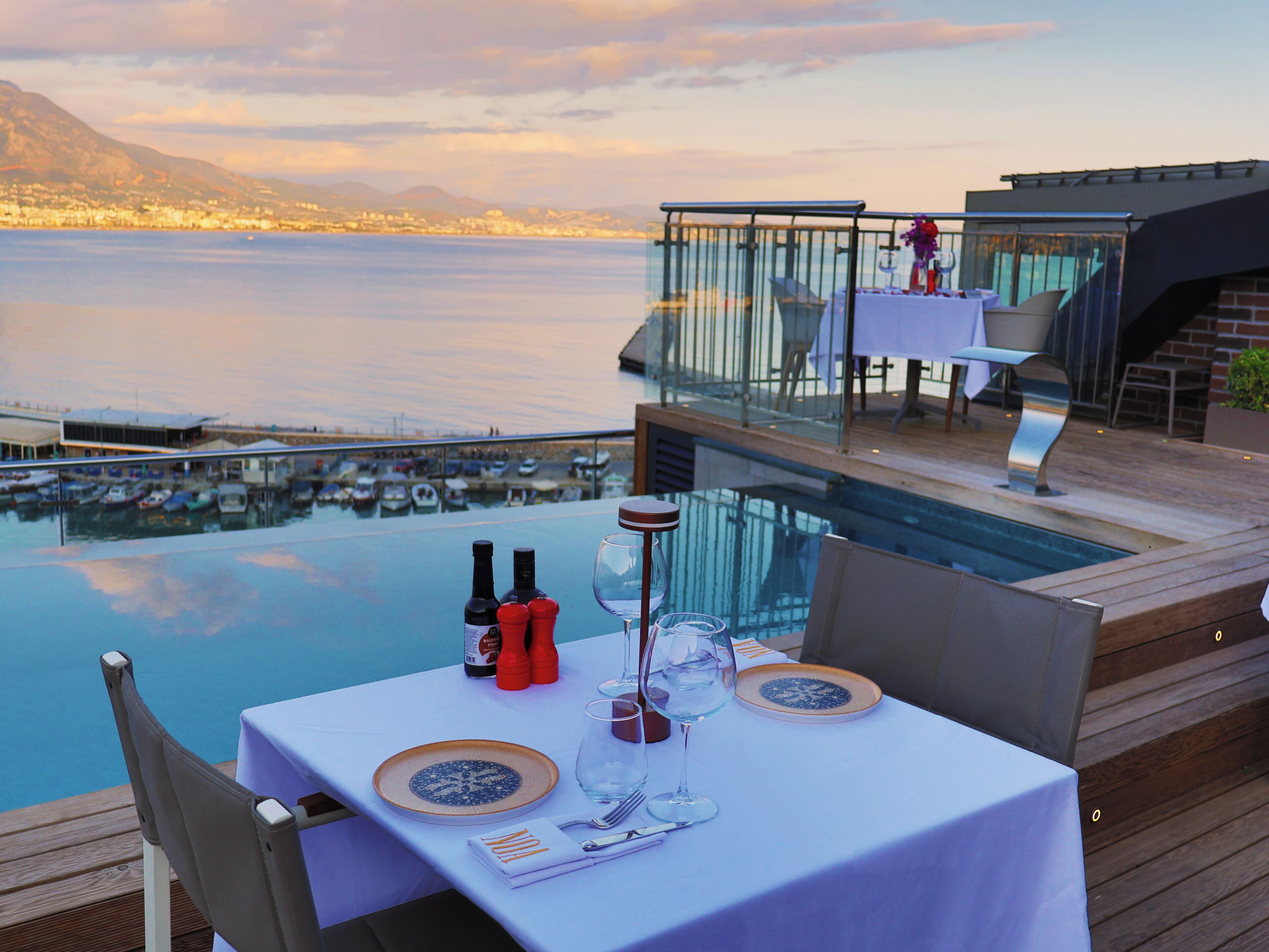 A dining table with a view of the sea and mountains, a glass railing, and a poolside setting.