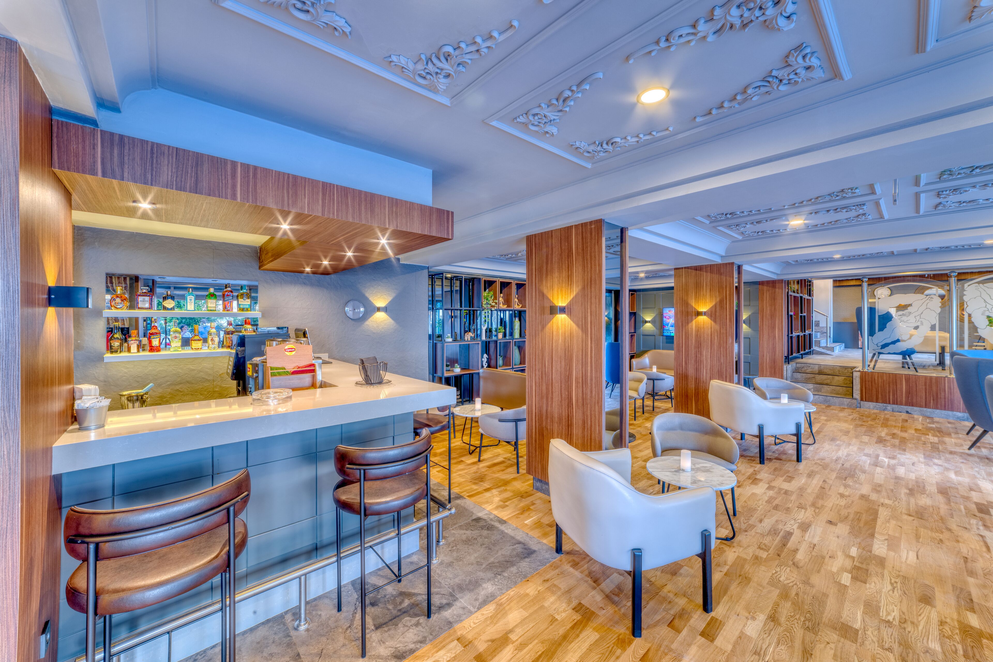 Zona de bar moderna con mostrador de madera, taburetes y un área de asientos con sillas y mesas blancas.
