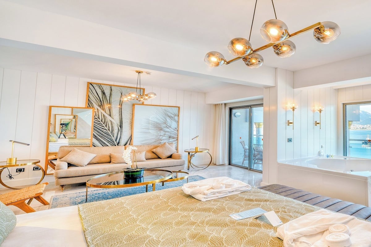 A modern hotel room with a large bed, a sitting area with a sofa and coffee table, a balcony with a view of the sea, and a bathtub.