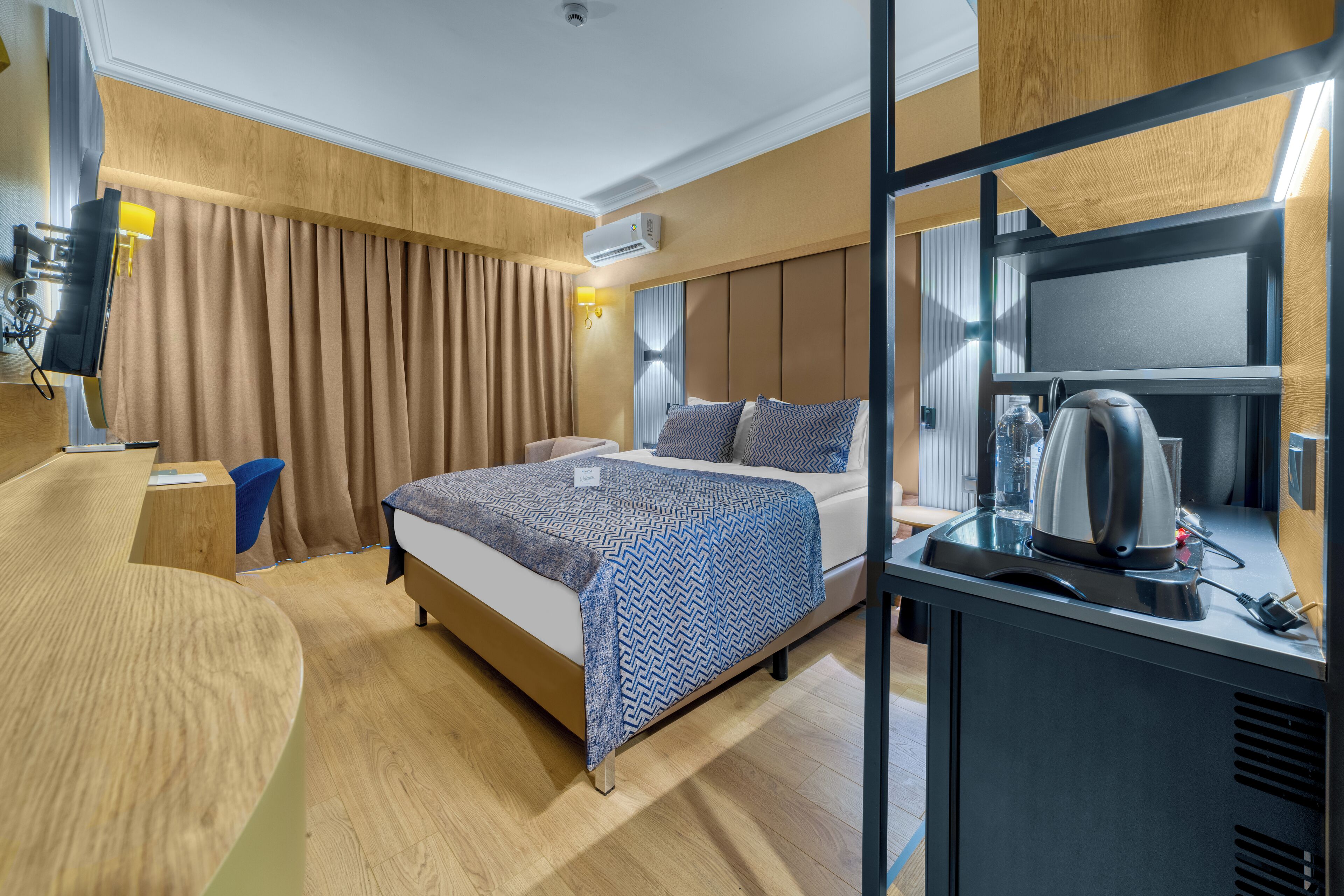A hotel room with a bed, bedside table, and a desk with a kettle.