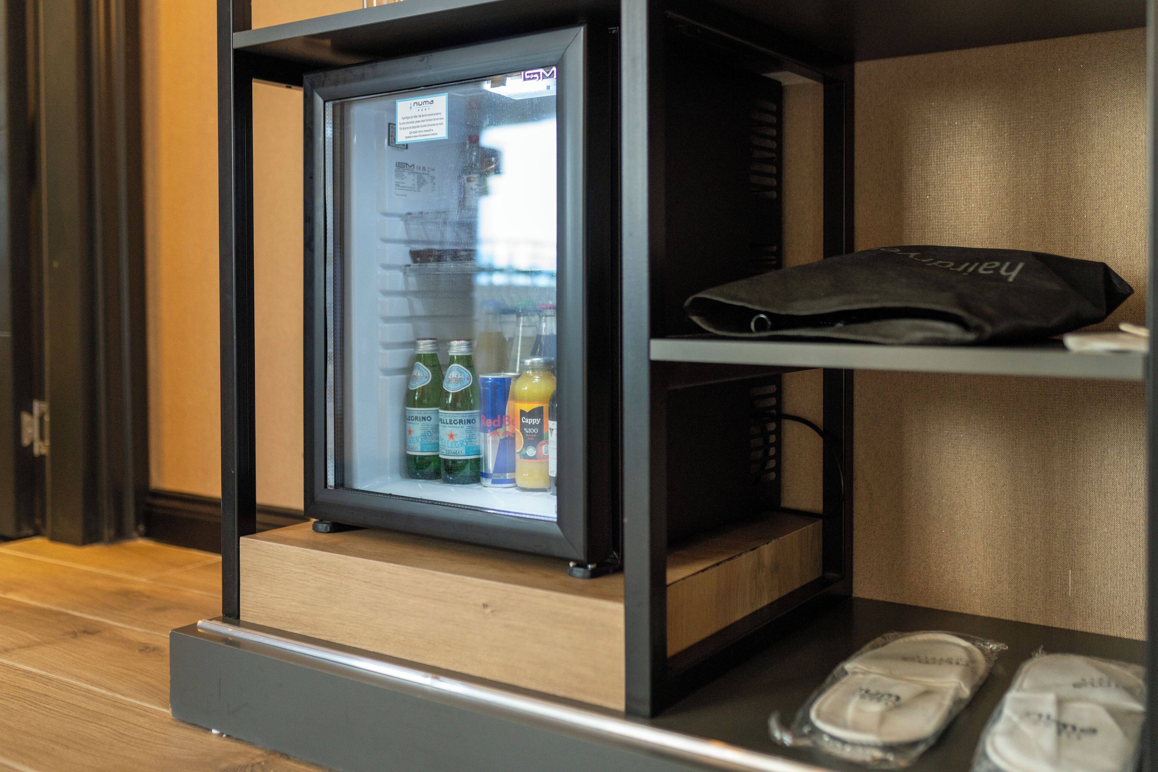 A minibar with a refrigerator containing bottled water, a juice, and a soda. There is a black bag on the shelf above the minibar.
