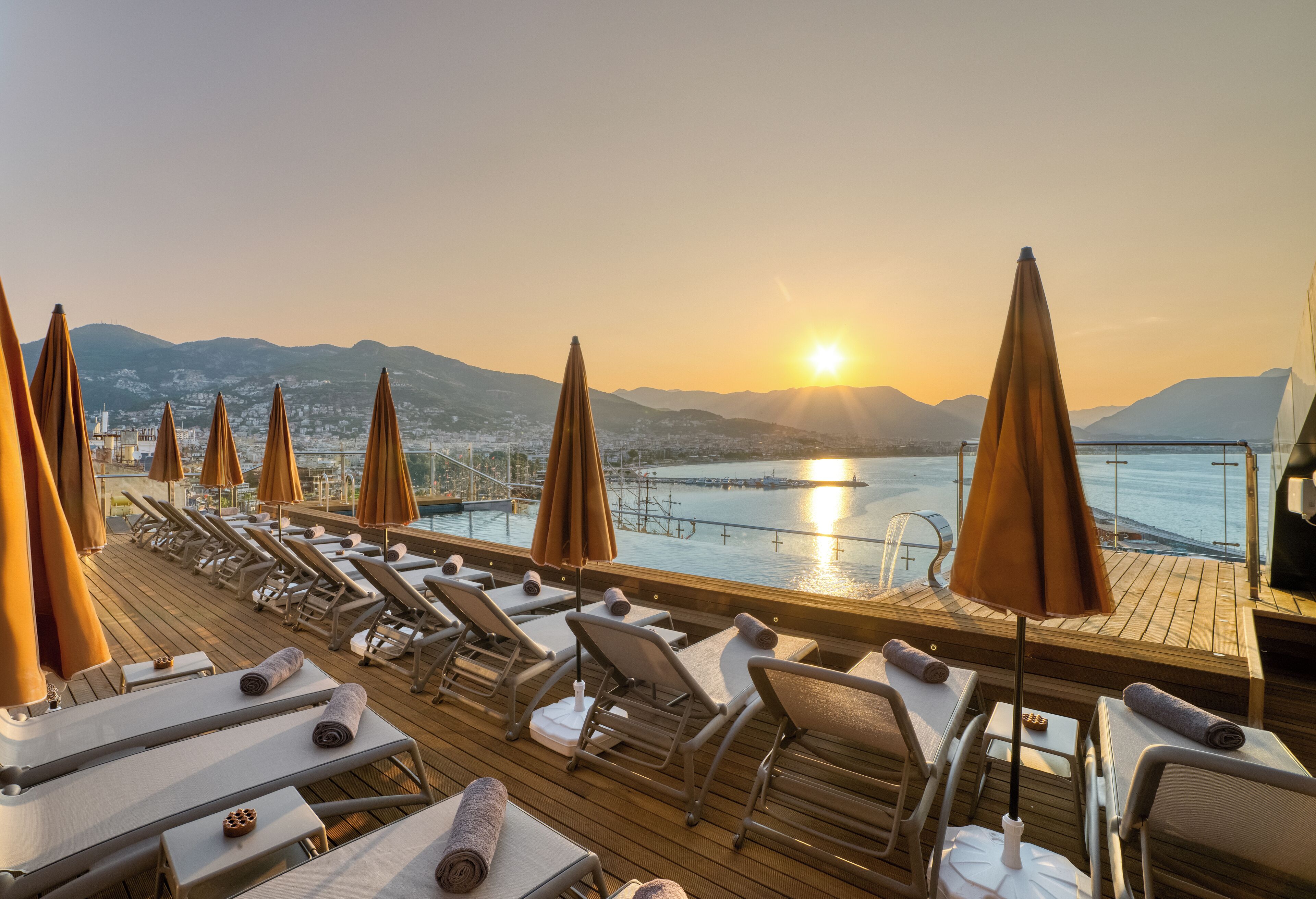 Una terraza iluminada por el sol con sillones de descanso y sombrillas, con vistas a un cuerpo de agua y montañas lejanas.