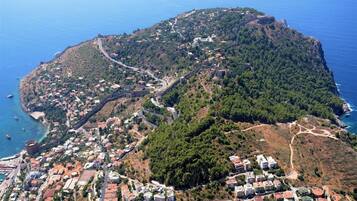An aerial view of a coastal town with a mix of residential buildings and natural terrain, including a prominent hill with a fort-like structure.