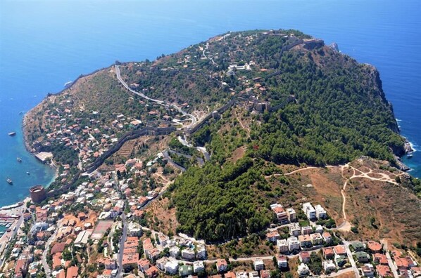 An aerial view of a coastal town with a mix of residential buildings and natural terrain, including a prominent hill with a fort-like structure. - Numa Port Hotel (Alanya)