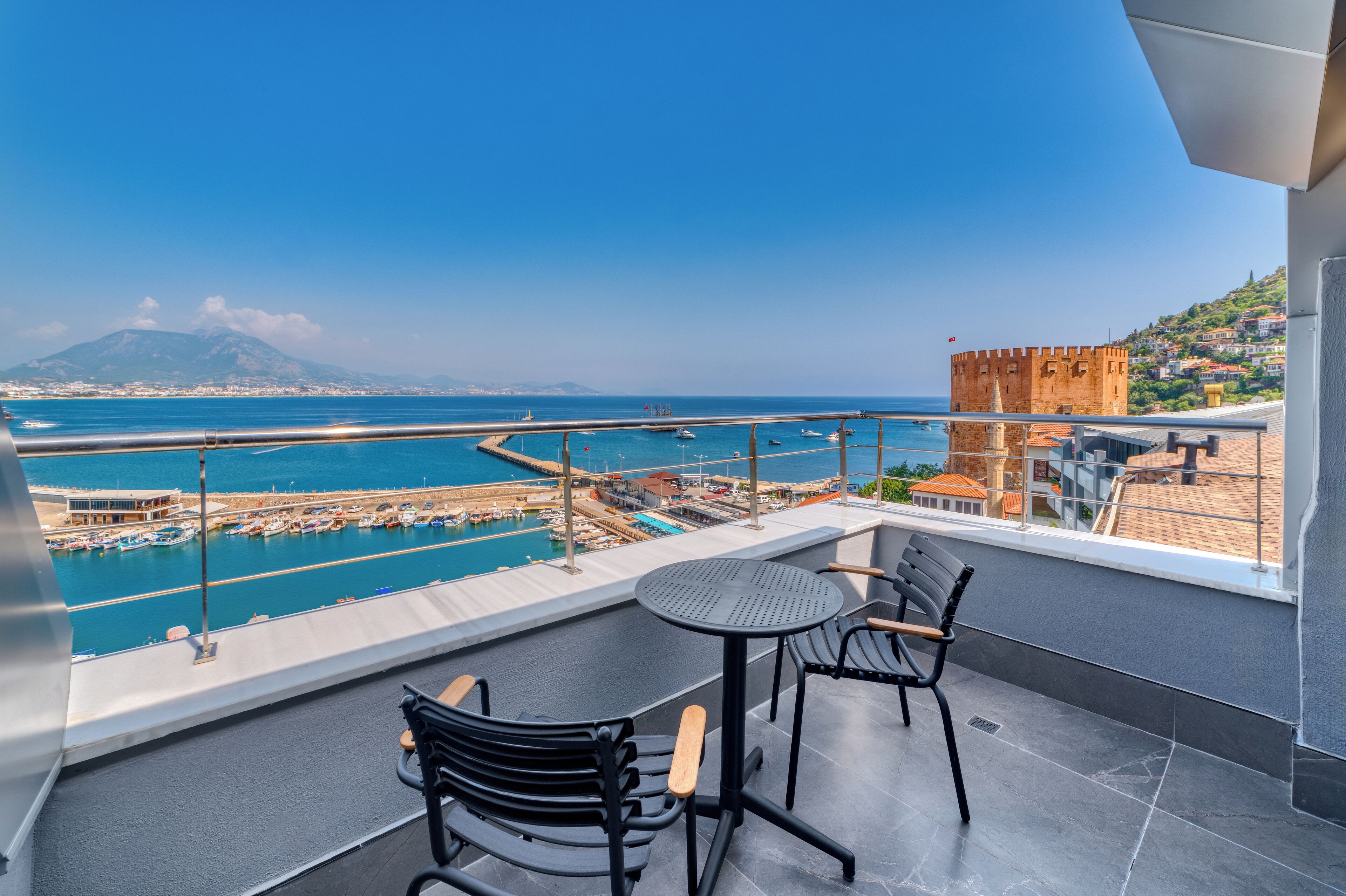 A balcony with two chairs and a table overlooking a marina with boats and a historic tower.