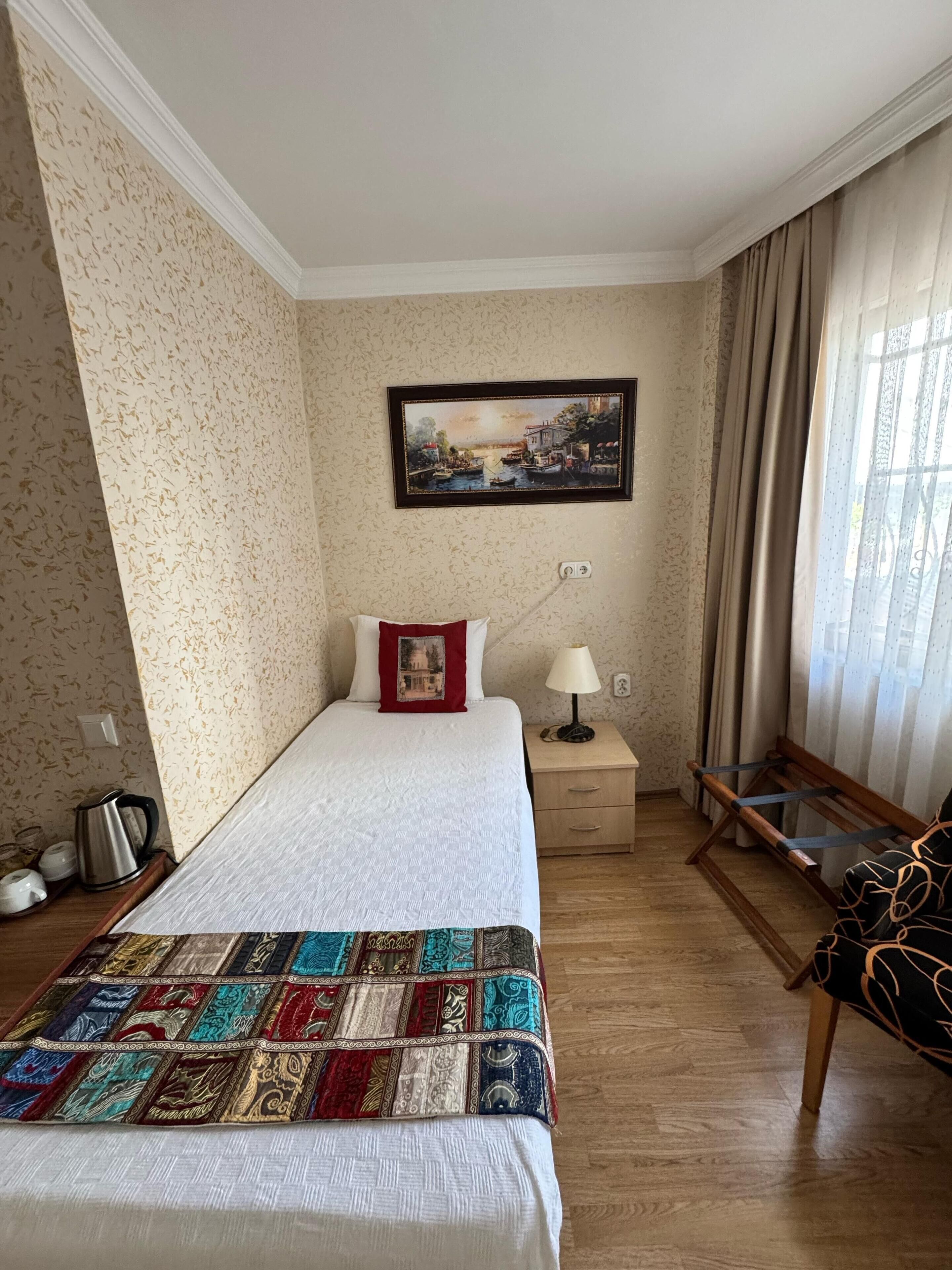 Una habitación de hotel ordenada con una cama, una mesita de noche, una lámpara, una silla y un cuadro en la pared.
