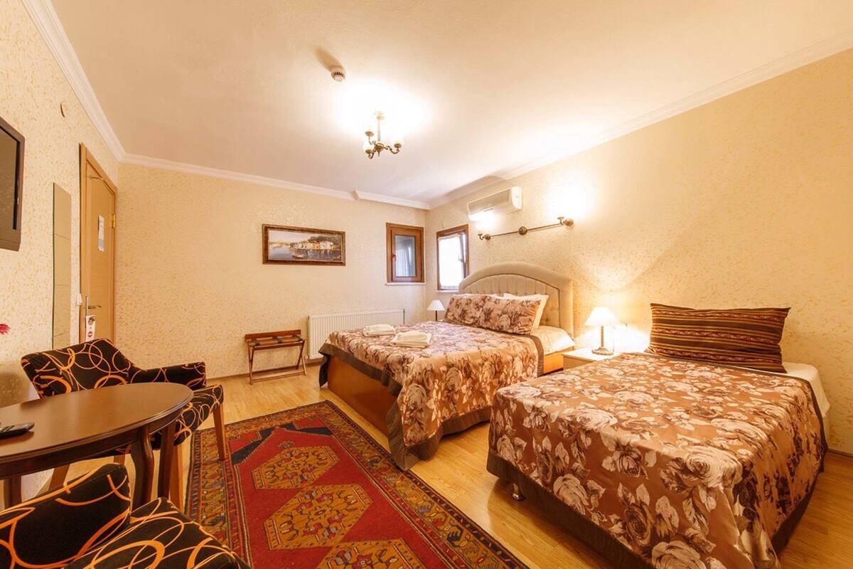 Habitación de hotel con dos camas, una mesa de comedor, sillas, un tapete rojo estampado y un cuadro en la pared.
