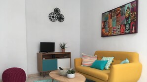Een moderne woonkamer met een gele bank, een ronde salontafel, een rode zitkussen, een televisie op een houten dressoir en een kunstwerk aan de muur.