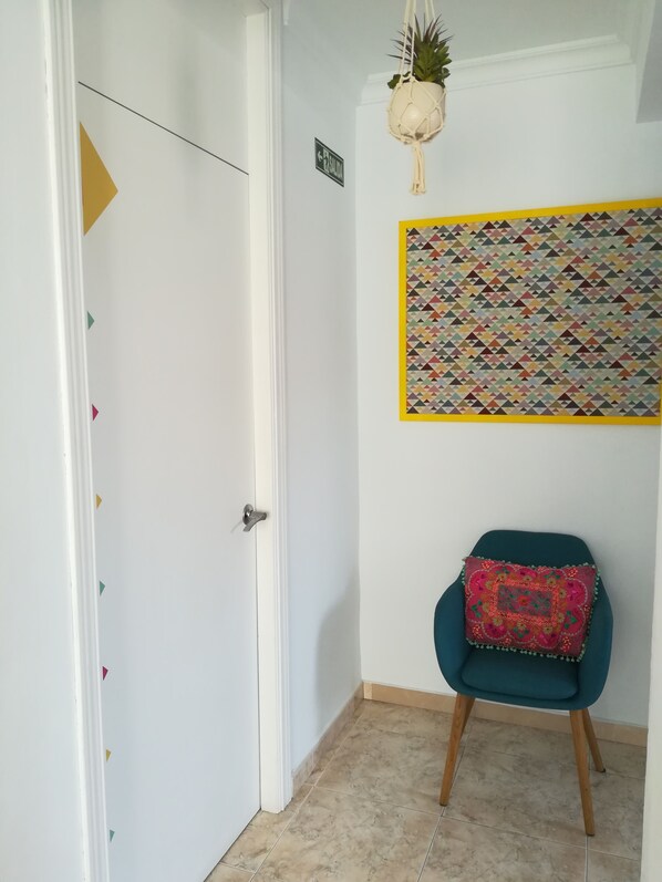 Un pasillo con una puerta blanca, una señalización, un arte mural geométrico de colores, un sillón azul turquesa con cojín estampado y una planta colgante.