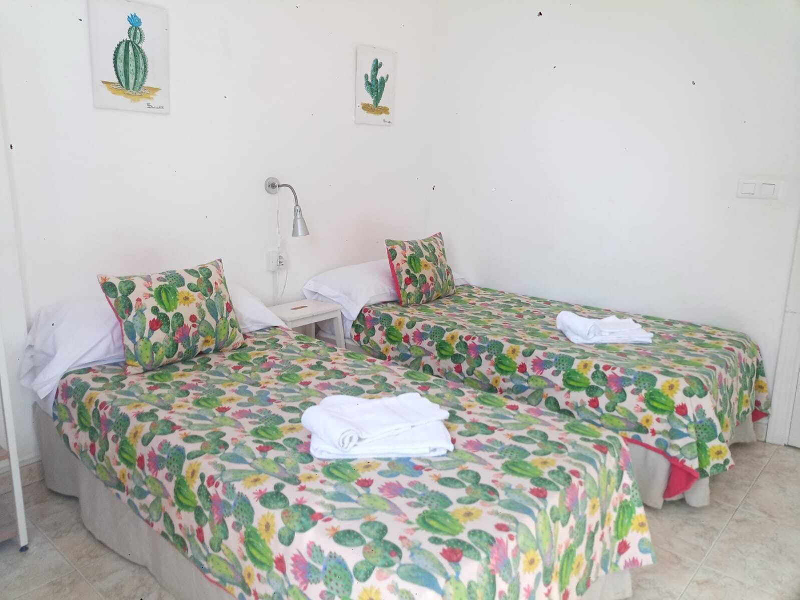 Habitación con dos camas, cada una con cubrecama con motivos de cactus, una mesita de noche pequeña y una lámpara.