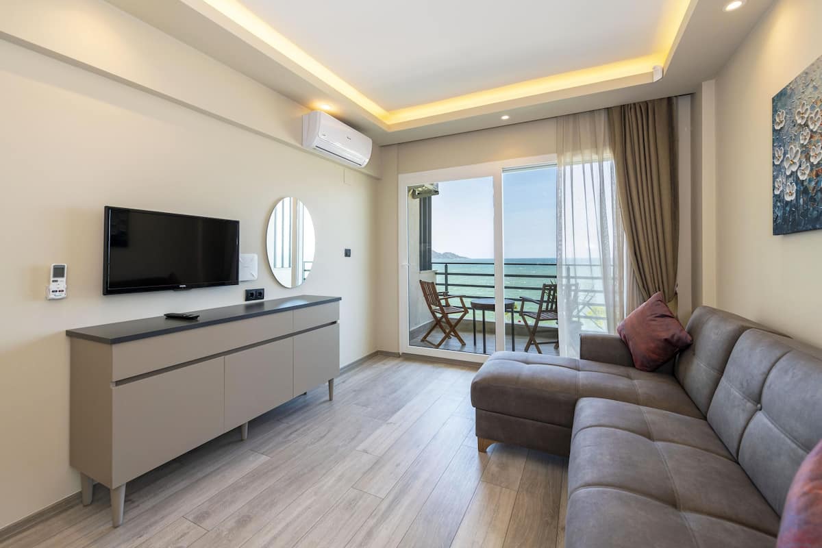A modern living room with a flat-screen TV, a sofa, a dining table with chairs, and a large window with a view of the sea.