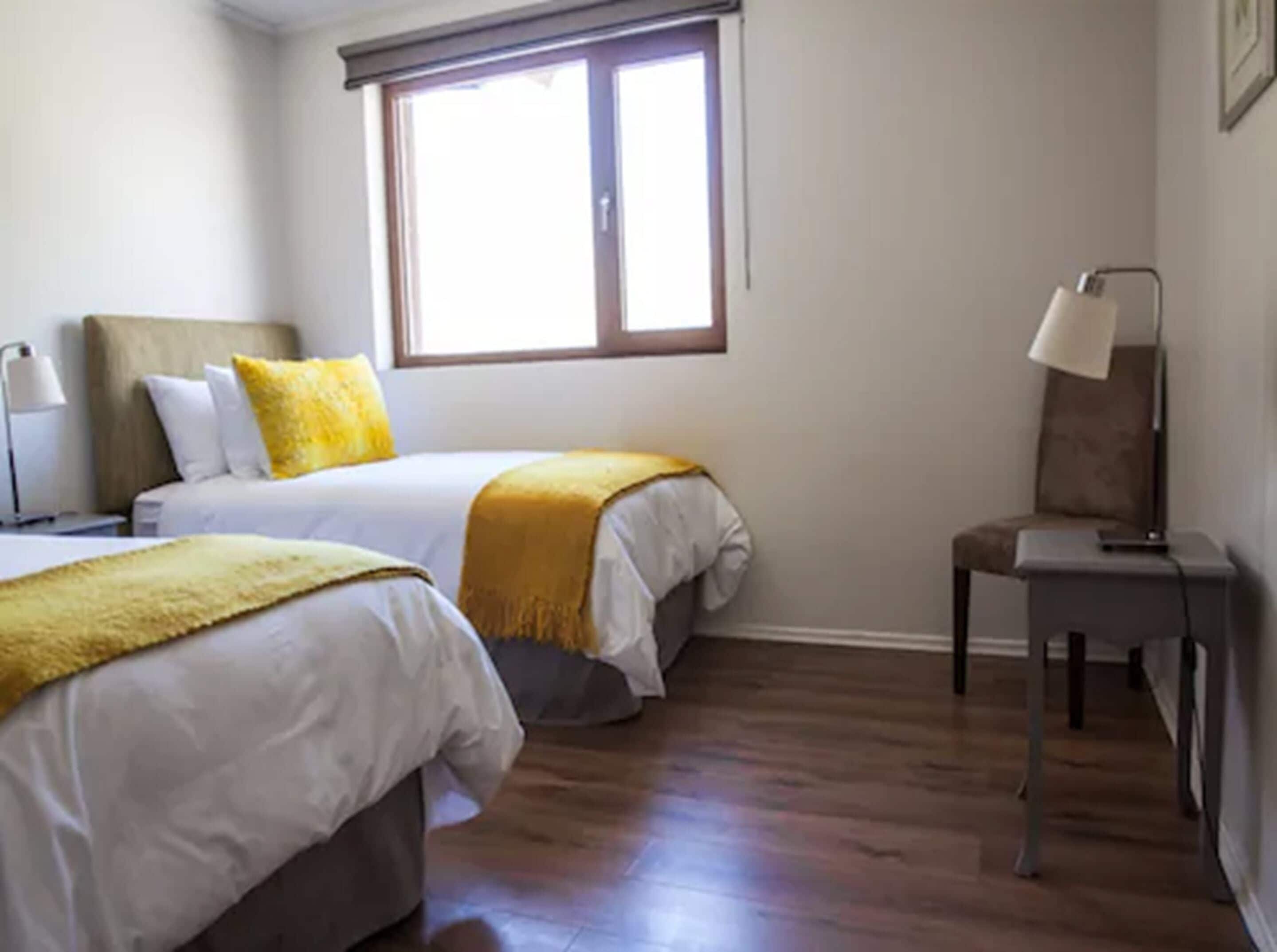 A bedroom with two beds, a window, a bedside table with a lamp, and a chair.