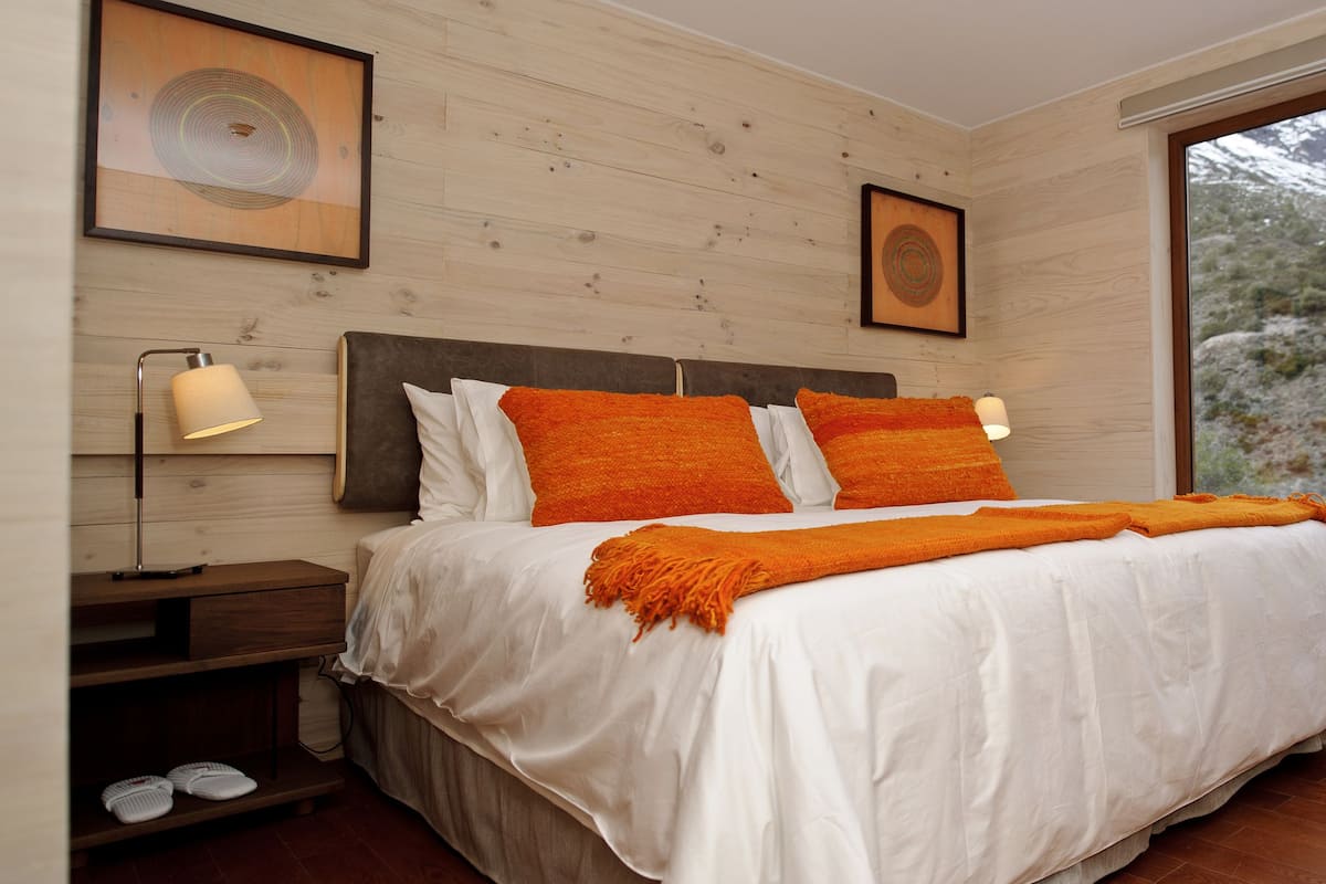A bedroom with a bed, two bedside lamps, a nightstand, and a window with a view of snow-capped mountains.