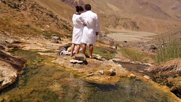 Two people in white robes standing by a clear, shallow stream in a mountainous area.