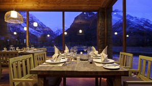 A dining table set for four with wooden chairs, large windows offering a view of snow-capped mountains, and pendant lighting. - Noi Puma Lodge (Machalí)