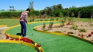 A person playing golf on a course with a green and yellow curvy path, surrounded by greenery and a chair in the background.