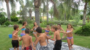 A group of children playing on a water slide in a grassy outdoor area with palm trees.