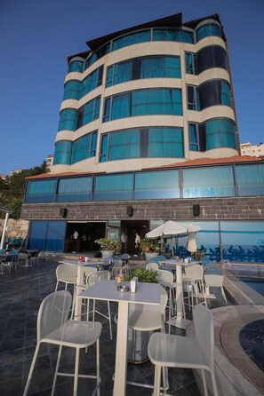 A modern building with a curved facade, large windows with blue curtains, outdoor seating with white tables and chairs, and a pool area. - Bay Lodge (Jounieh)