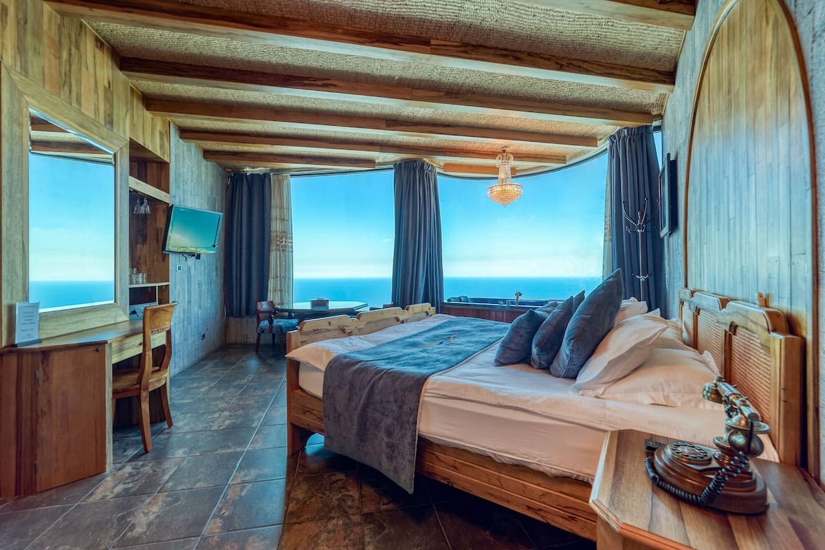 A bedroom with a large bed, wooden headboard, a desk, and a chair. There is a television on the wall and a view of the ocean through the windows.