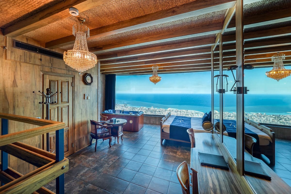 A high-ceilinged room with a city view, wooden flooring, a bed, a desk, and a seating area with a view.