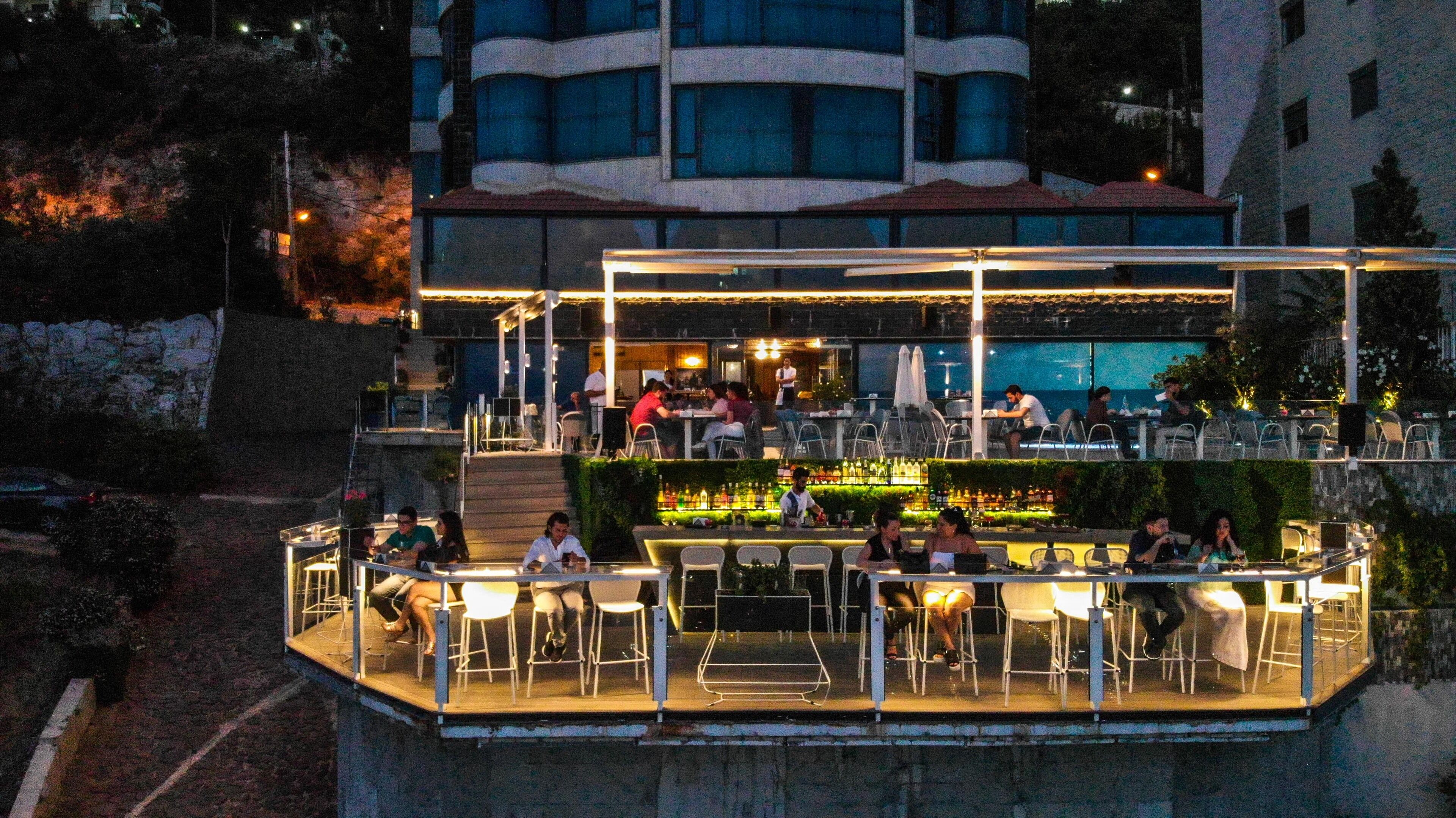 A rooftop bar with outdoor seating, a pool in the background, and a stone facade.
