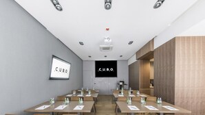 Moderni kokoushuone, jossa on useita pöytiä ja tuoleja, projektorikangas ja suuri näyttö, jossa näkyy teksti ”CUBO HOTEL”.