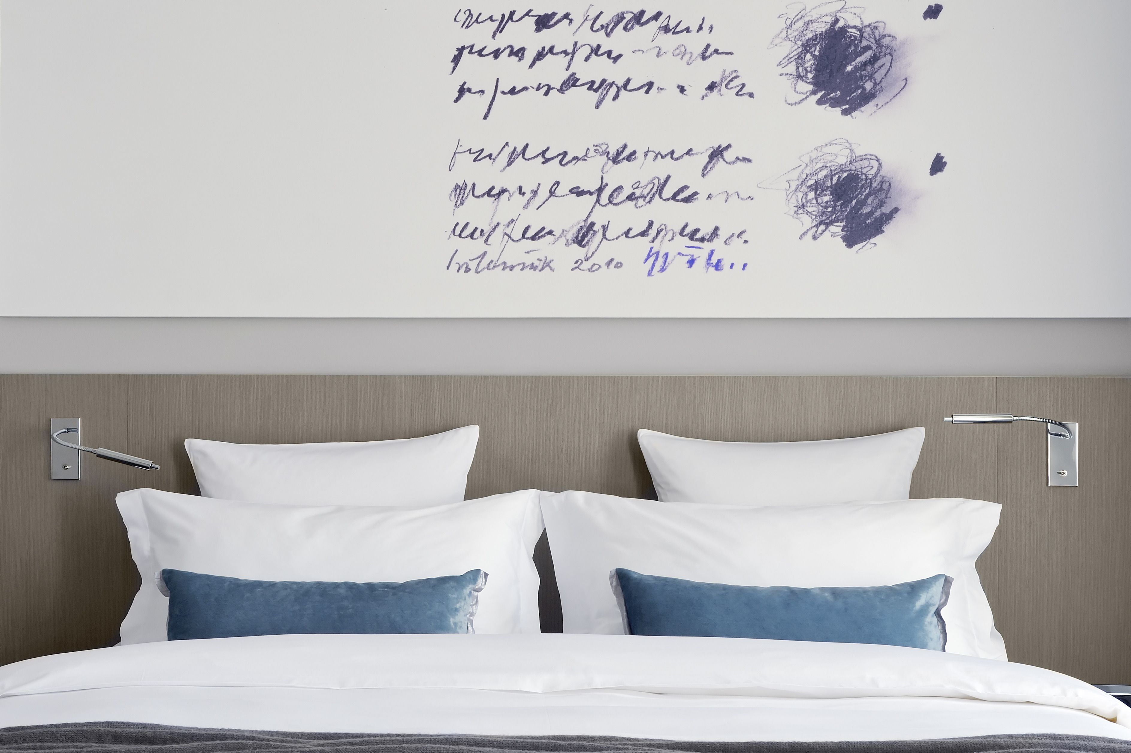 Une chambre d’hôtel moderne avec un lit soigneusement fait, des oreillers blancs et bleus, et un mur orné d’œuvres d’art abstraites et d’inscriptions.