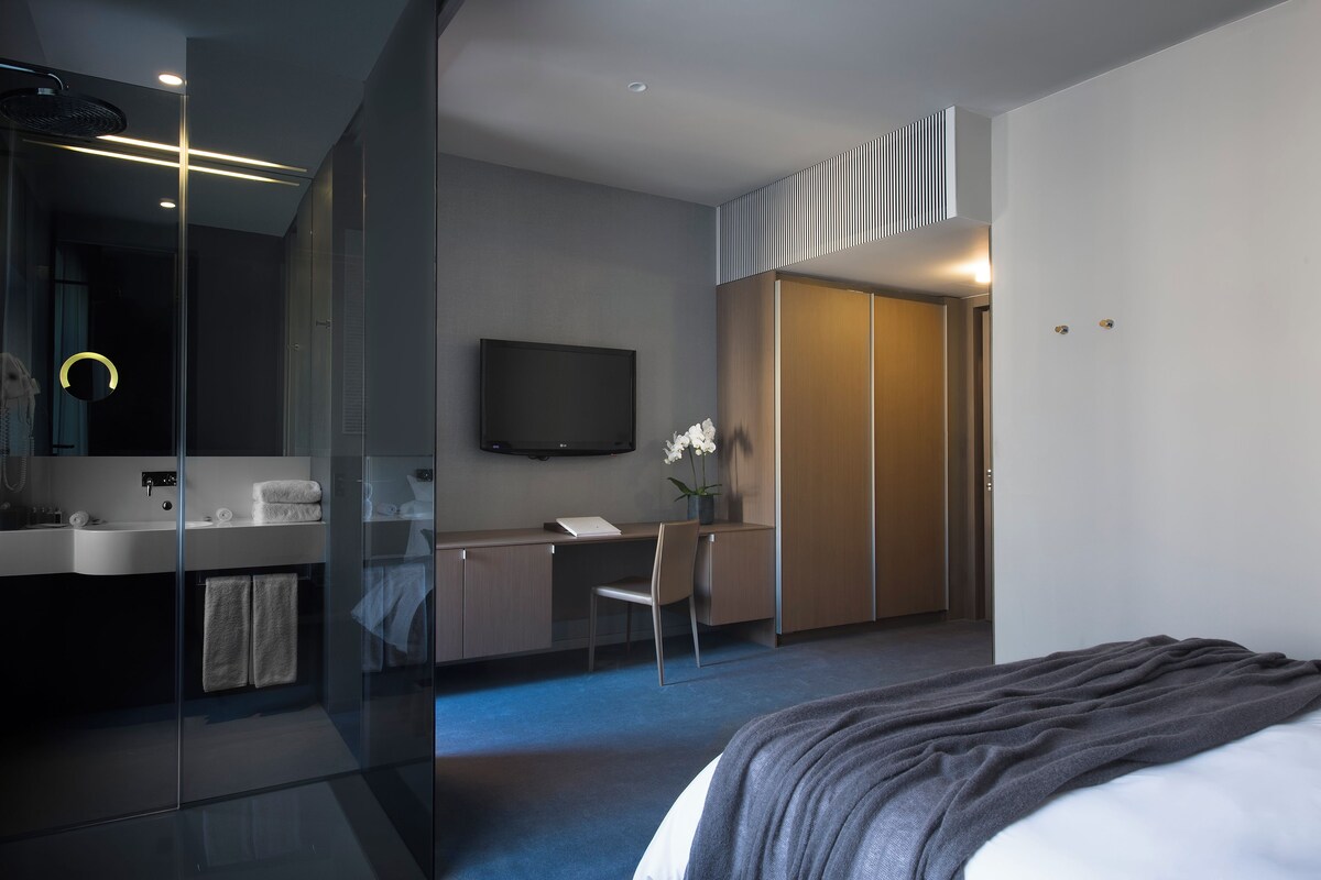 Moderni hotellihuone, jossa on lasiseinällä varustettu suihku, työpöytä tuolilla, taulutelevisio ja harmaalla peitolla varustettu sänky.