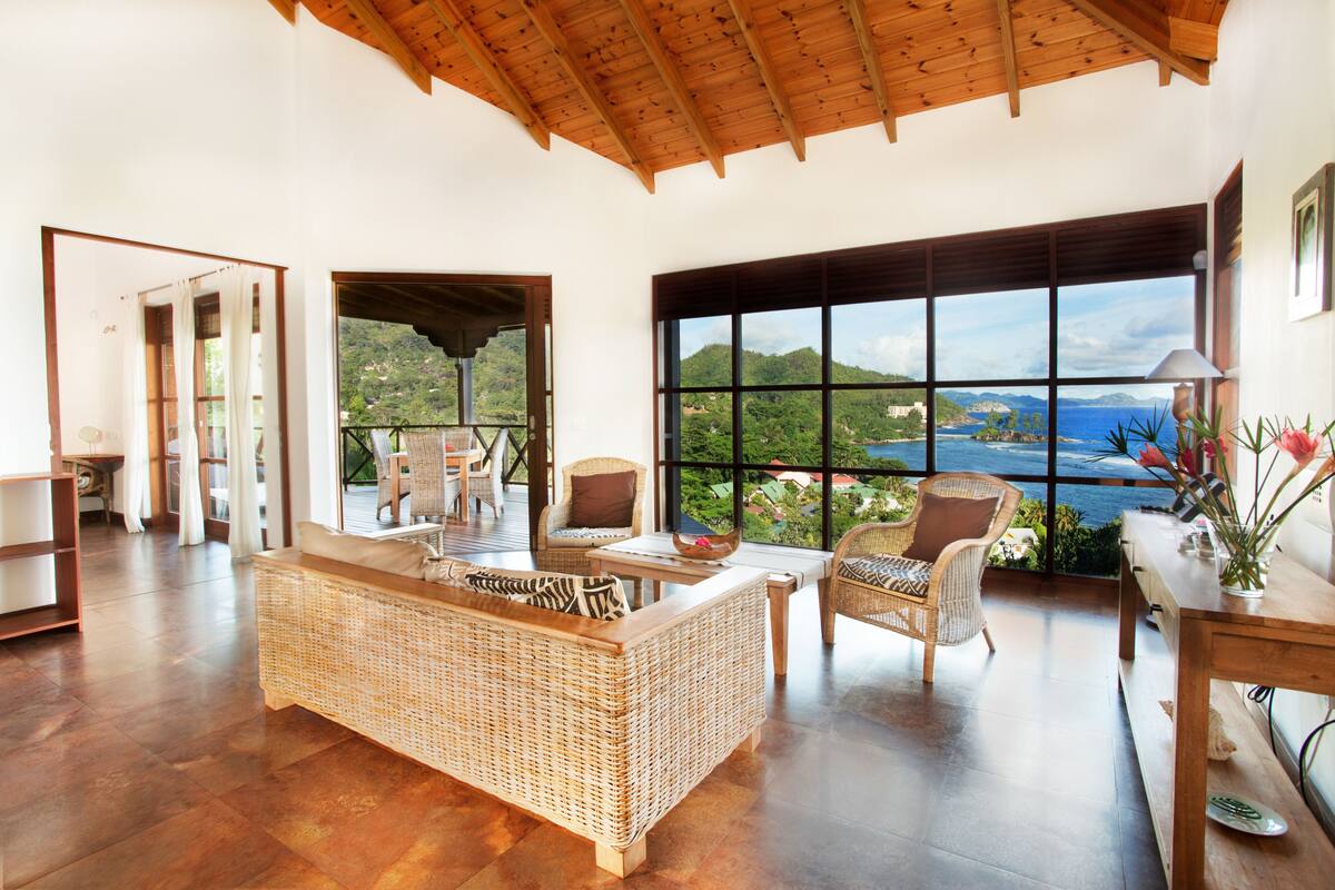 Un vaste espace de vie comprenant un grand canapé en osier, des fauteuils en osier et une table en bois. De grandes fenêtres offrent une vue sur un paysage montagneux et la mer.