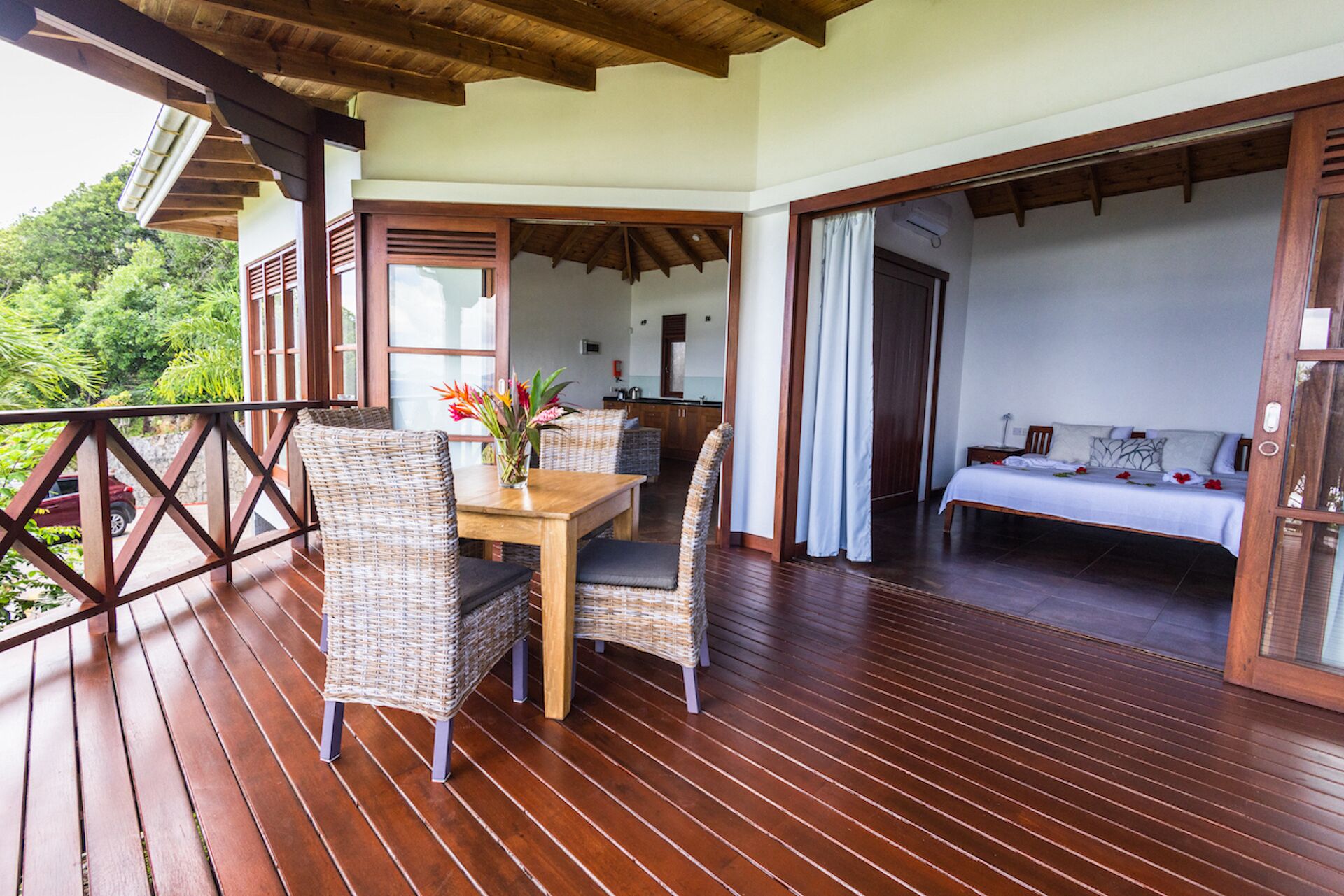 A wooden deck with wicker furniture, a dining table, and a bed in the adjacent room.