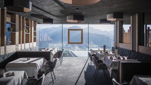 A modern dining area with tables and chairs, a large window with a mountain view, and a unique ceiling design. - Hotel Hubertus (Valdaora)