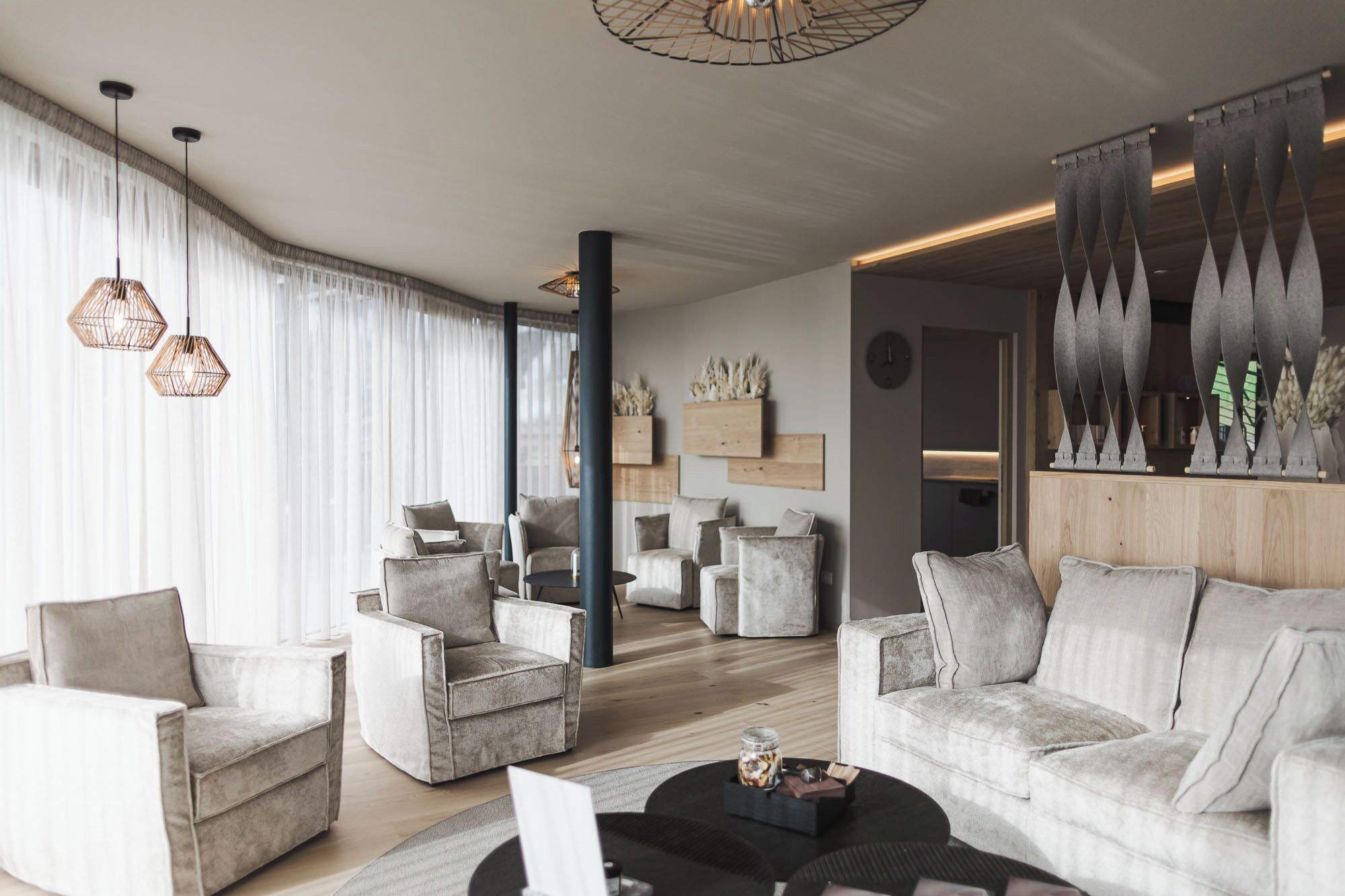 A modern living room with a sofa, armchairs, and a coffee table. There are pendant lights, a wooden accent wall, and a view into another room.