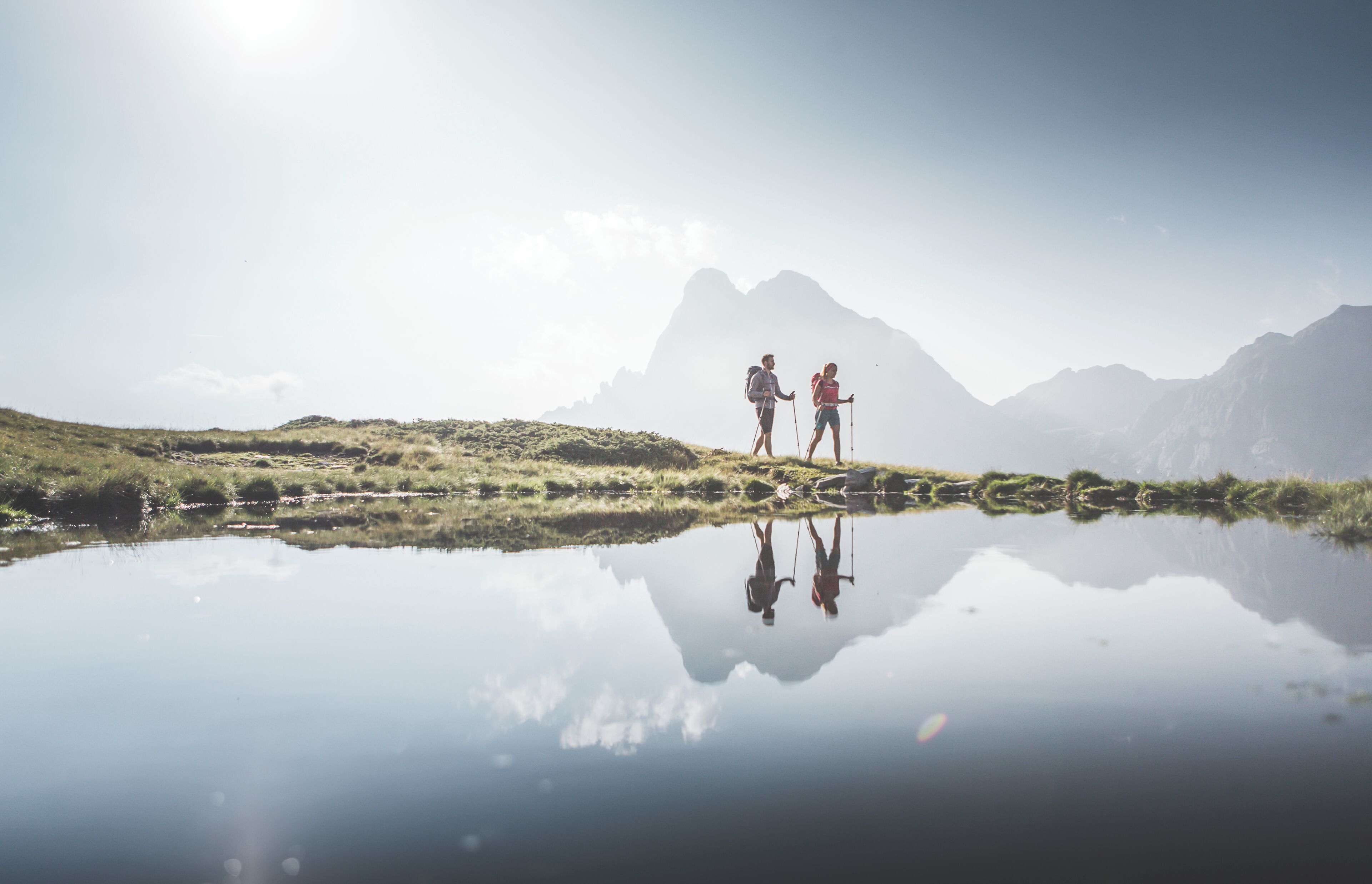Dos personas caminando por senderos cerca de un cuerpo de agua que refleja el paisaje, con montañas al fondo.